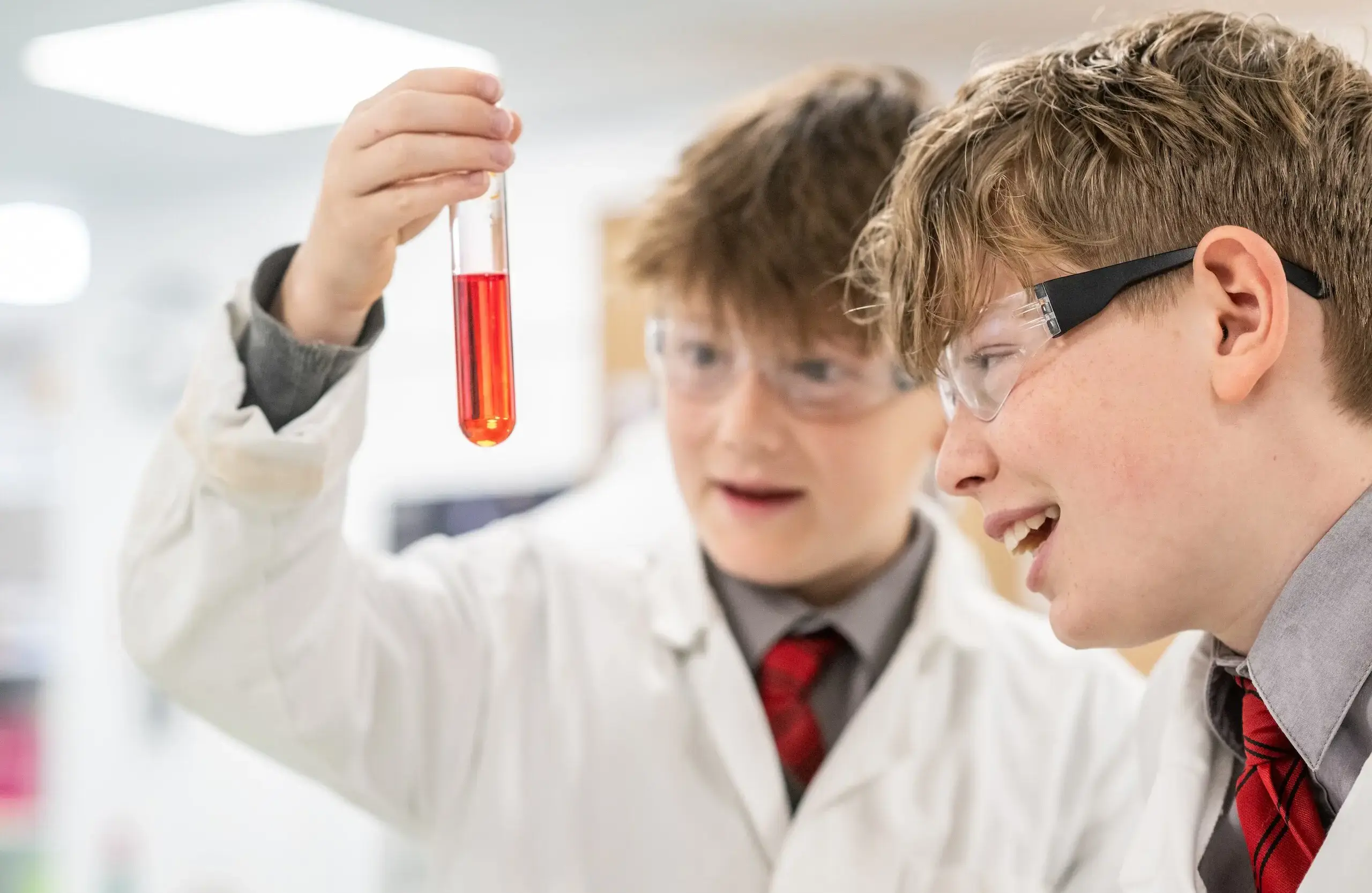 Two students, both wearing lab coats and safety goggles, examine a vial full of deep orange liquid