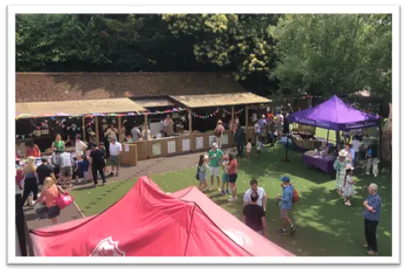 People gathered outdoors at a summer fair with stalls, tents, and decorations.